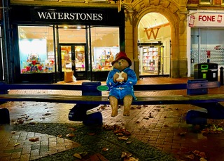 A photo of Paddington bear sitting on a bench in Broad St, Reading, eating a marmalade sandwich.